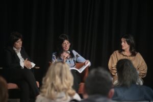Tre donne sedute su una scena durante un evento, una di loro parla in un microfono.